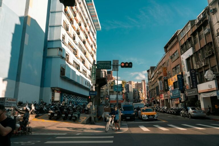 City street scene with modern buildings, traffic, and pedestrians under a clear summer sky.