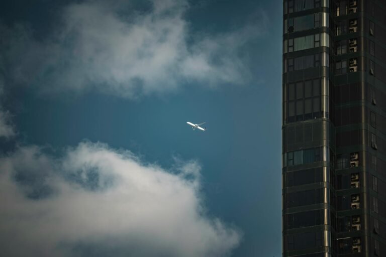 A lone airplane flies through a partly cloudy sky beside a modern skyscraper in Bangkok, Thailand.