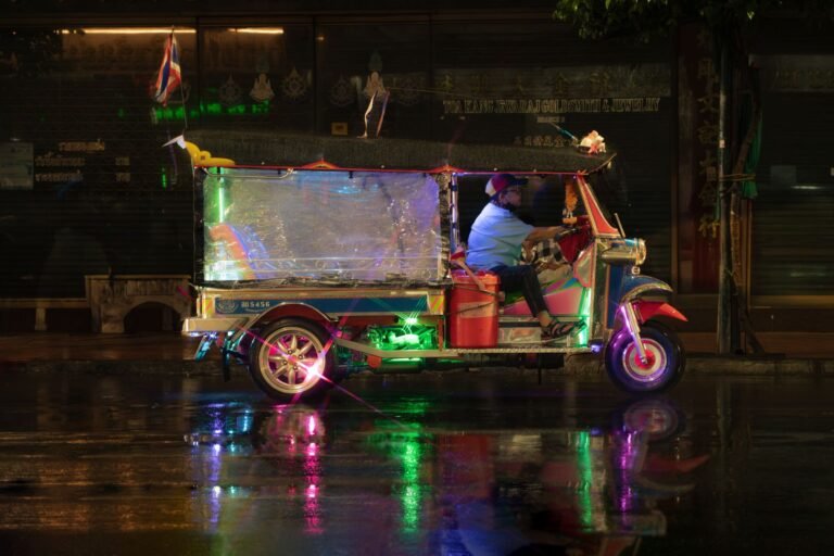 A vibrant tuk-tuk drives through a rainy Bangkok street, reflecting colorful lights.